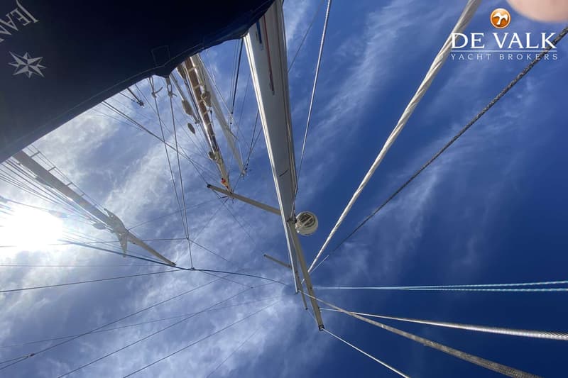 De foto toont een mast en tuigage van een zeilboot, gezien vanuit een laag perspectief tegen een blauwe lucht met wolken.