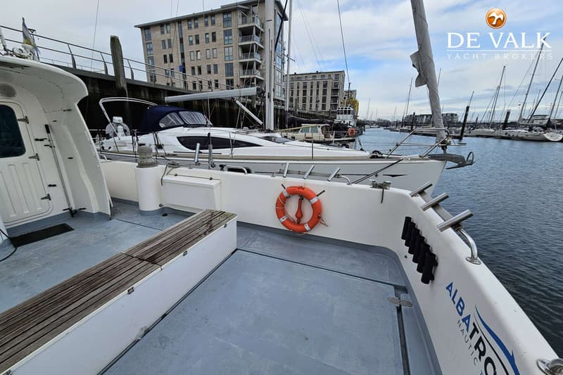 Een witte boot met de naam "ALBATROS NAUTIC" op de zijkant ligt in een haven met andere boten en gebouwen op de achtergrond.