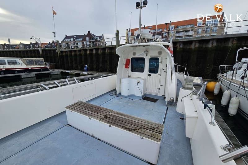 Een witte boot met een houten bankje op het dek, aangemeerd in een haven met gebouwen op de achtergrond. — Waypoint 34