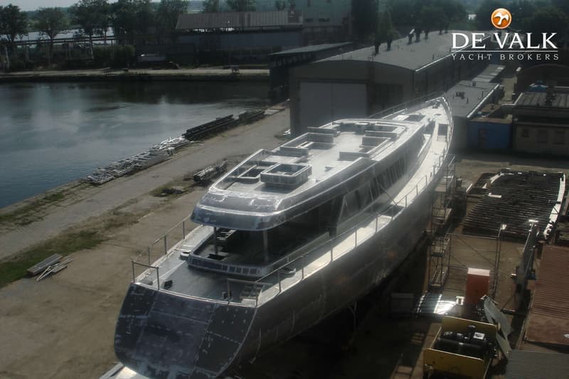 Een onafgebouwde, grijze boot staat op de kade naast een waterweg, met gebouwen en bomen op de achtergrond.