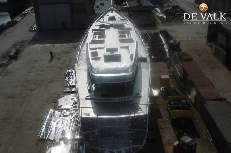 Een onafgewerkte, grijze boot met een bovenbouw en relingen, gezien van bovenaf op een werf.