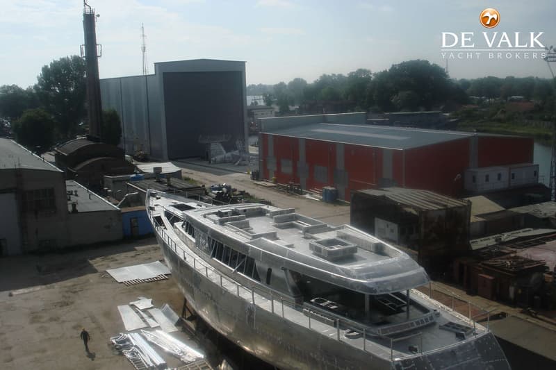 Een onafgewerkte, zilverkleurige boot staat op een scheepswerf met gebouwen en een schoorsteen op de achtergrond.