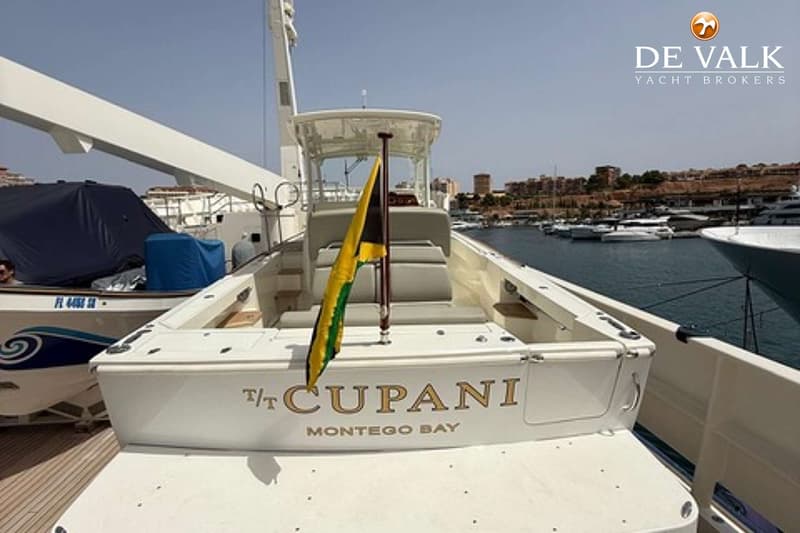 Een witte boot met de naam "T/T CUPANI MONTEGO BAY" op de spiegel, met een Jamaicaanse vlag en een haven op de achtergrond.