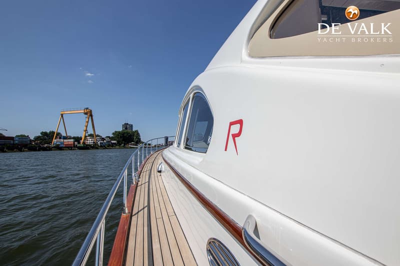 Een witte boot met een houten dek en een rode letter "R" op de zijkant vaart op een rivier. — Rapsody 55