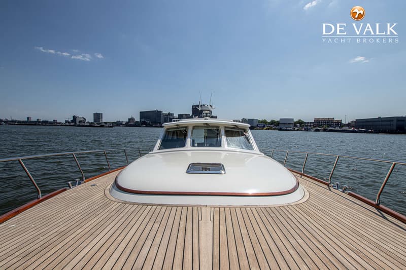Een witte motorboot met een teakhouten dek vaart op een waterweg met een stad op de achtergrond. — Rapsody 55