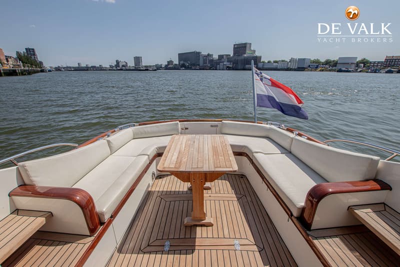 Een boot met een houten dek, een houten tafel, witte kussens en een Nederlandse vlag, varend op water met een stad op de…