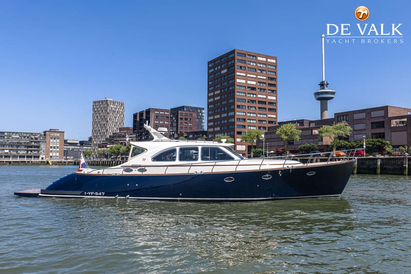 Een donkerblauwe motorboot met witte opbouw vaart op het water, met gebouwen en een toren op de achtergrond. — Rapsody 55