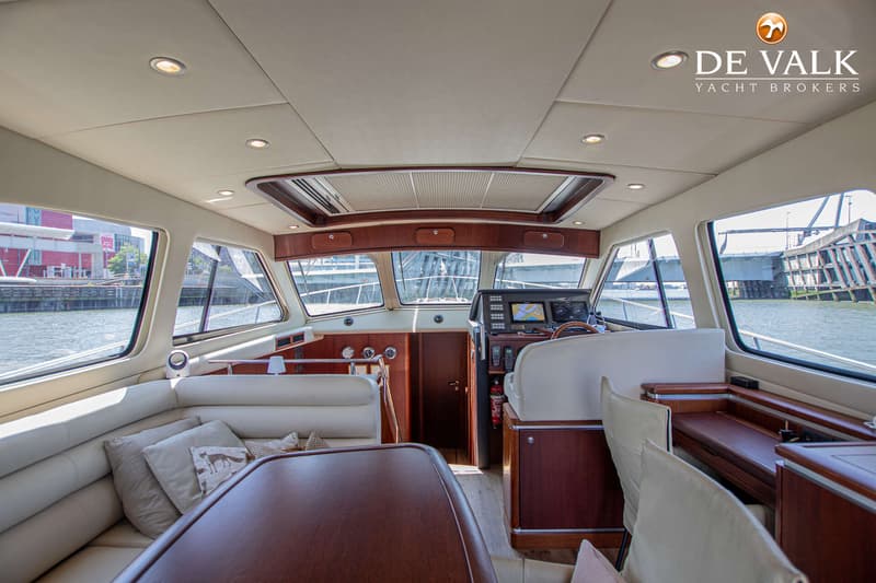 De foto toont het interieur van een boot met een stuurstand, zitbanken, een tafel en uitzicht op water en gebouwen.