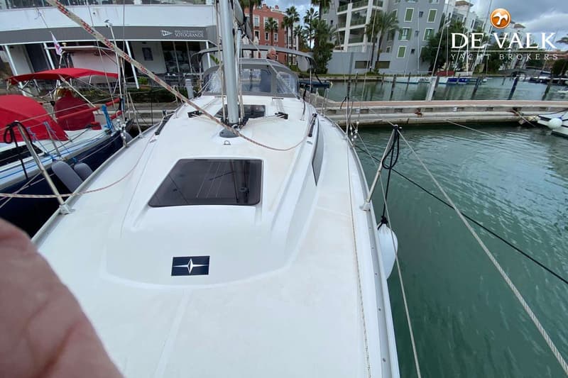Een witte zeilboot ligt aangemeerd in een haven, met een man in de kuip en gebouwen op de achtergrond. — Bavaria 34 Cruiser