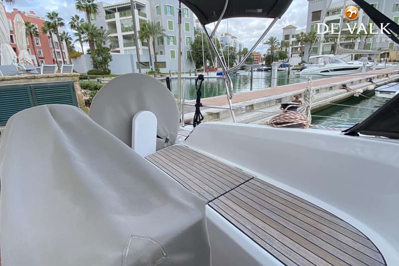 Een boot met een teakdek en afdekhoezen ligt in een jachthaven met gebouwen en palmbomen op de achtergrond.