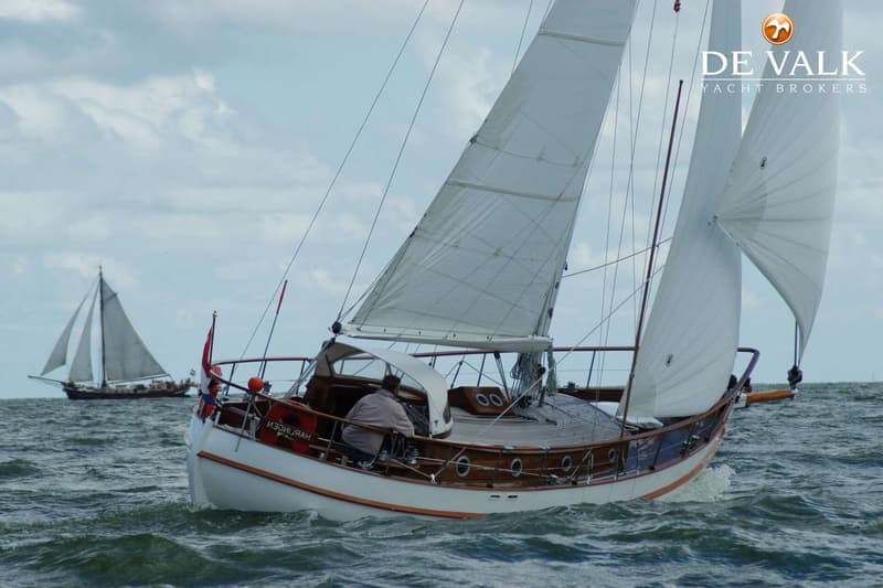 Een zeilboot met witte romp en houten details vaart op golvend water, met een persoon aan het roer en een andere zeilboot…
