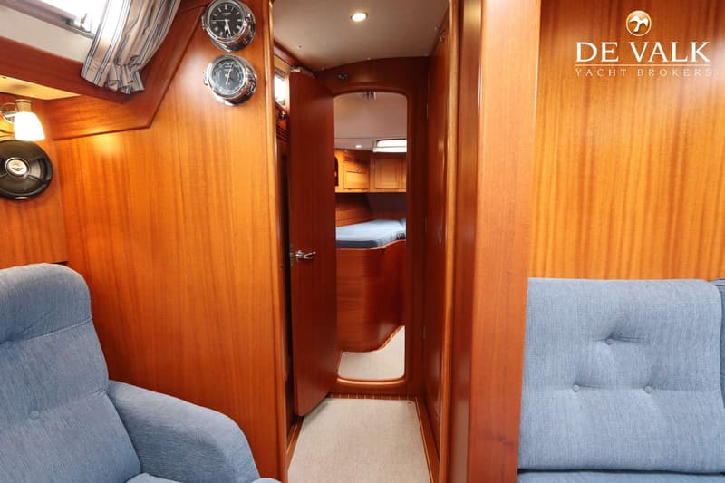 Een interieur van een boot met houten wanden, twee blauwe stoffen stoelen, en een open deur naar een slaapcabine.