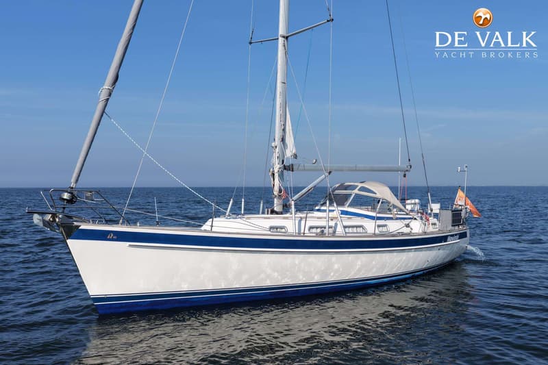 Een witte zeilboot met blauwe strepen en een grijze buiskap vaart op kalm water onder een helderblauwe lucht.