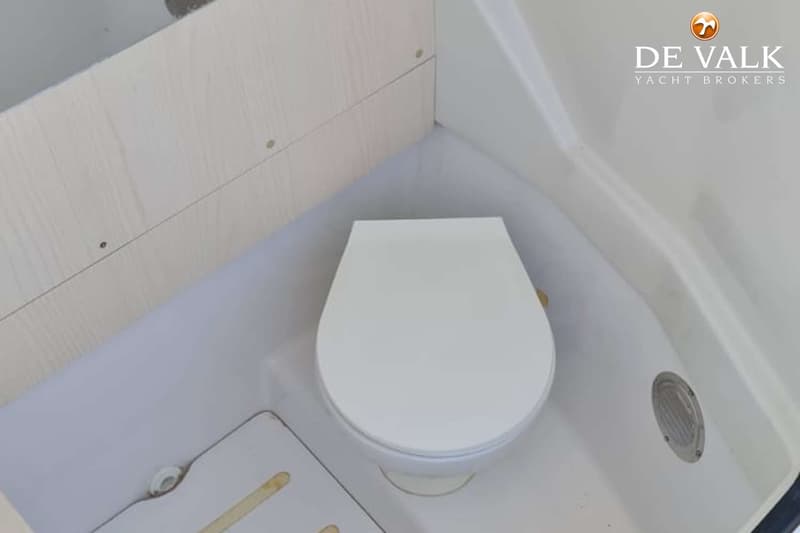 Een witte boottoilet met gesloten deksel staat in een hoek, met een houten wand en een ventilatierooster.