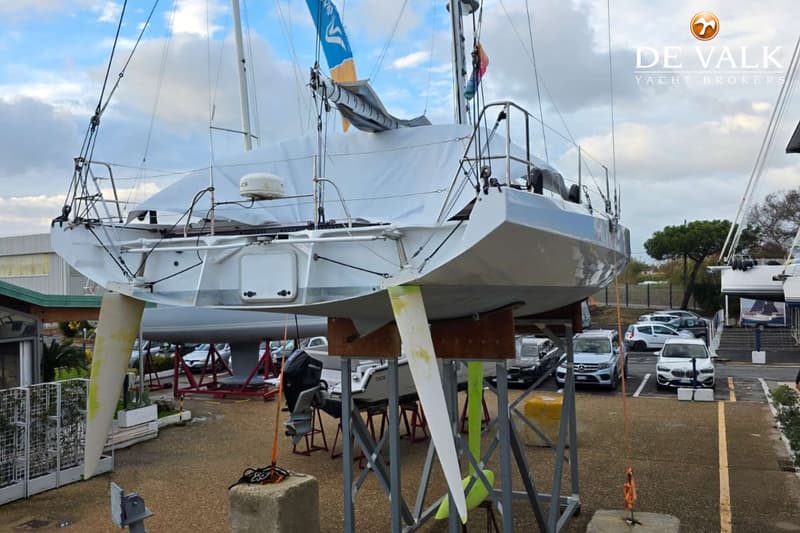 Een witte zeilboot staat op een trailer op de wal, met een witte hoes over het dek en een blauw-geel zeil opgerold.