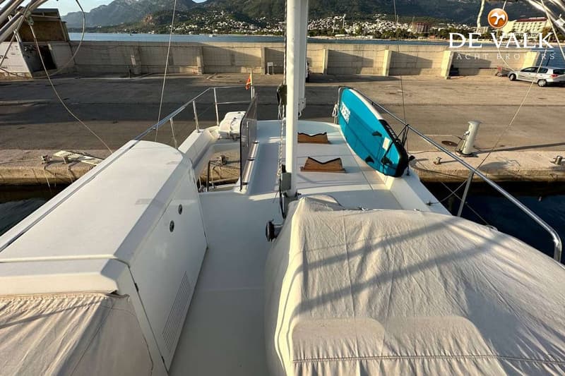 De foto toont het dek van een boot met een blauw paddleboard, een mast en afgedekte objecten, met een kade en bergen op de…