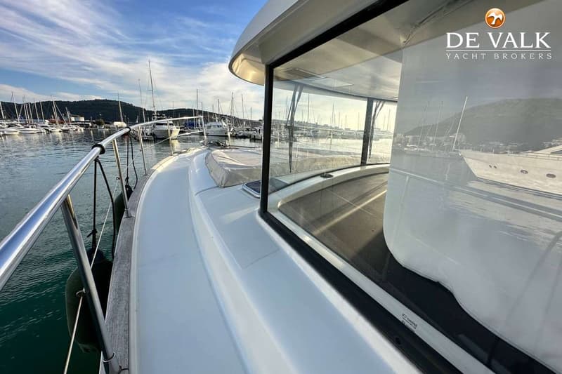 Een witte boot met een reling en stootwillen ligt in een haven met andere boten en bergen op de achtergrond.