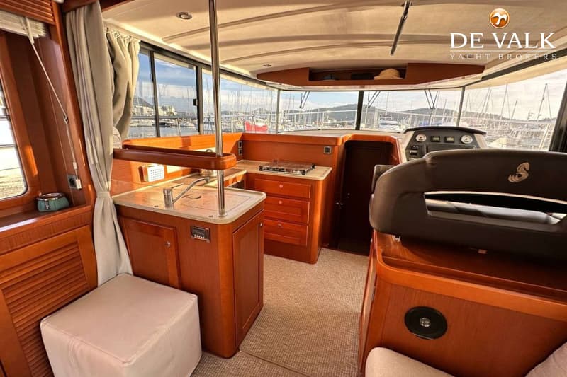 De foto toont het interieur van een boot met een kombuis, stuurstand en uitzicht op een haven vol boten.