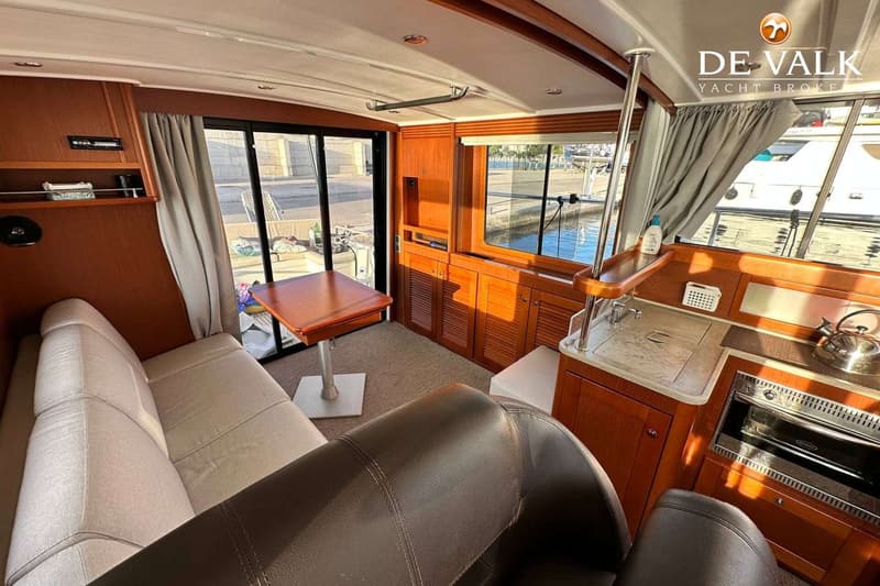 De foto toont het interieur van een boot met een zithoek, tafel, keukenblok en ramen met uitzicht op water en een kade.