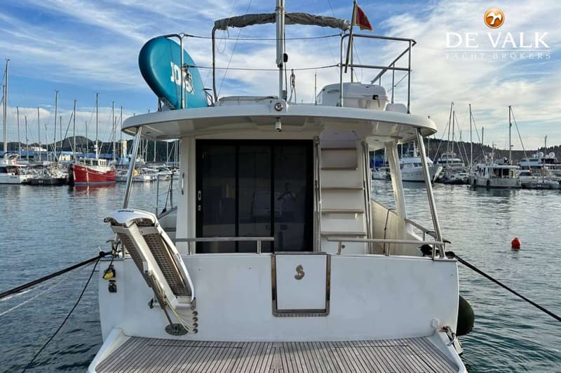 Een witte boot met een teakhouten zwemplateau en een blauw surfplank op het dak, afgemeerd in een haven.