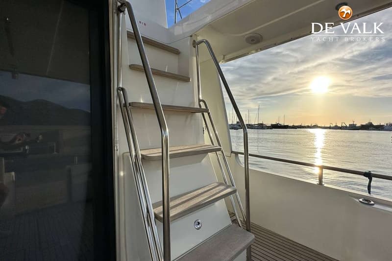 Een trap met houten treden en metalen leuningen leidt naar een bovendek, met uitzicht op water en boten bij zonsondergang.