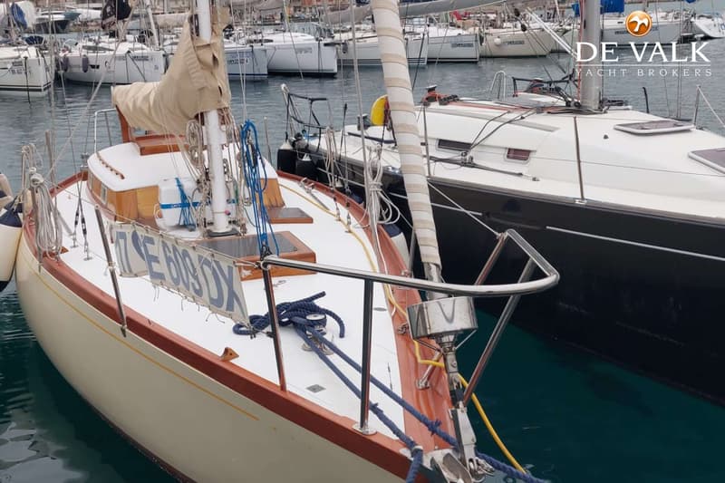 Een zeilboot met een crèmekleurige romp en houten details ligt in een haven, met andere boten op de achtergrond.