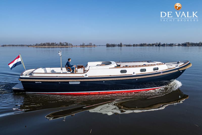 Een donkerblauwe boot met witte opbouw en rode onderkant vaart op een kalm wateroppervlak met een man aan het roer.