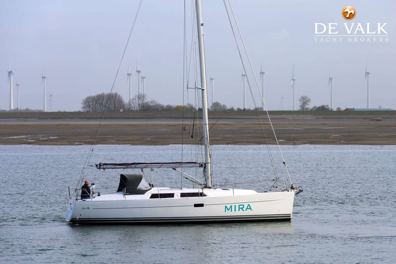 Een witte zeilboot met de naam "MIRA" in turquoise letters vaart op water met op de achtergrond windmolens en een landschap.