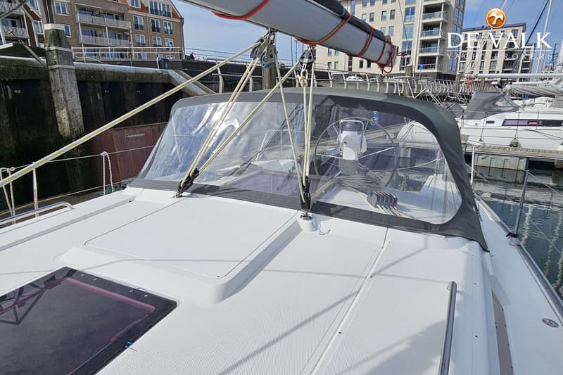 Een witte boot met een grijze buiskap en een opgerold zeil ligt in een haven met gebouwen op de achtergrond. — Hanse 375