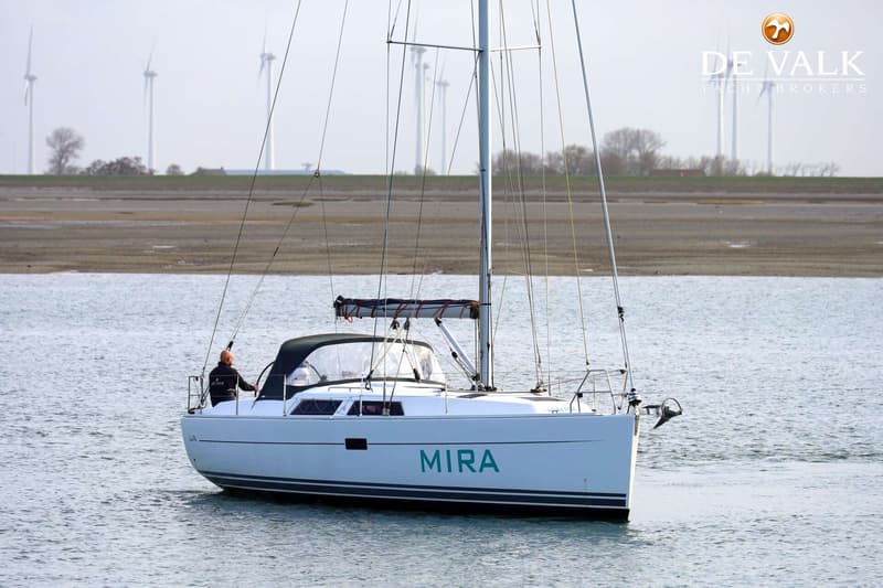 Een witte zeilboot met de naam "MIRA" in groene letters vaart op water, met een persoon aan het roer en windmolens op de…