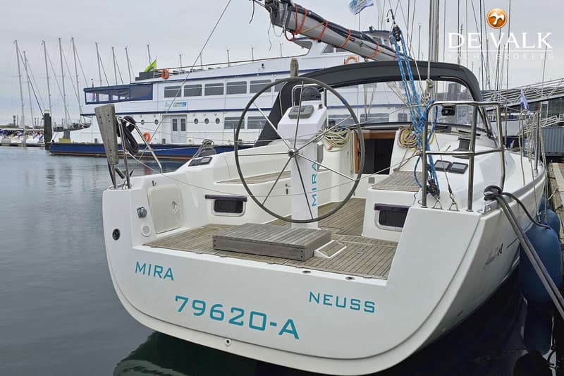 Een witte zeilboot met een teakhouten dek en een stuurwiel, afgemeerd in een haven met andere boten op de achtergrond.