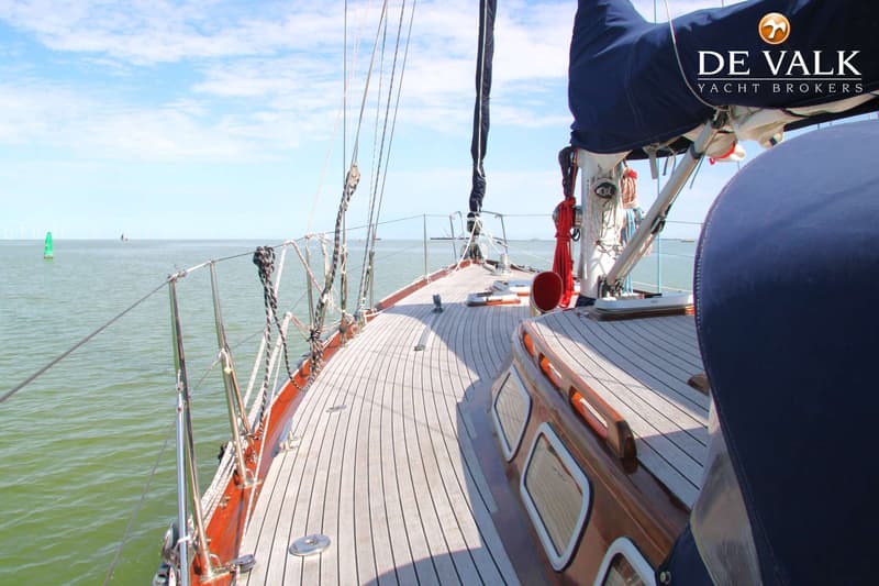 Een zeilboot met een houten dek en een blauw zeil op het water, met een logo van "DE VALK YACHT BROKERS". — Huisman 45