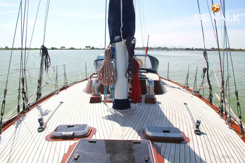 Een vooraanzicht van een zeilboot met een houten dek, touwen en een mast, varend op rustig water. — Huisman 45