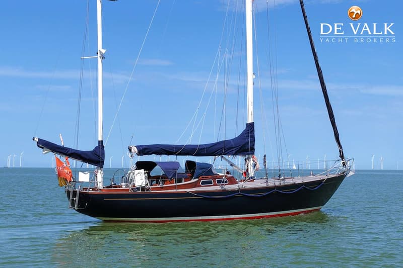 Een donkerblauwe zeilboot met een rode waterlijn en twee masten ligt op het water met windmolens op de achtergrond.