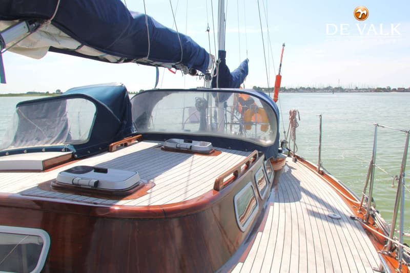 Een zeilboot met een houten dek en een blauwe buiskap, liggend op het water met een kustlijn op de achtergrond. — Huisman 45