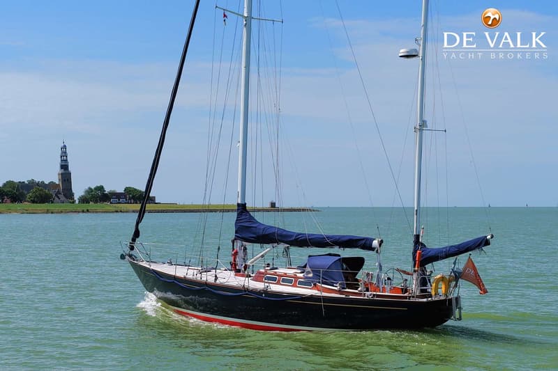 Een donkerblauwe zeilboot met rode streep vaart op groen water, met een kerktoren op de achtergrond. — Huisman 45