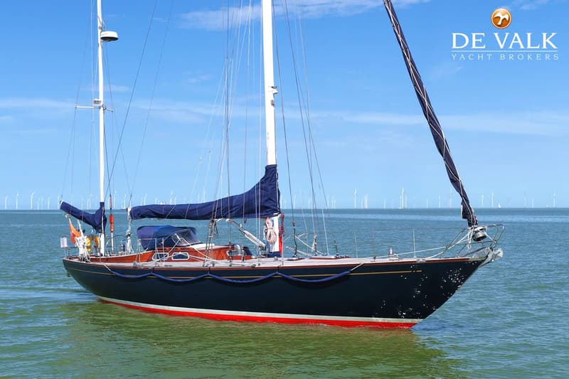 Een donkerblauwe zeilboot met rode en witte strepen onder de waterlijn, drijvend op groen water onder een blauwe lucht.