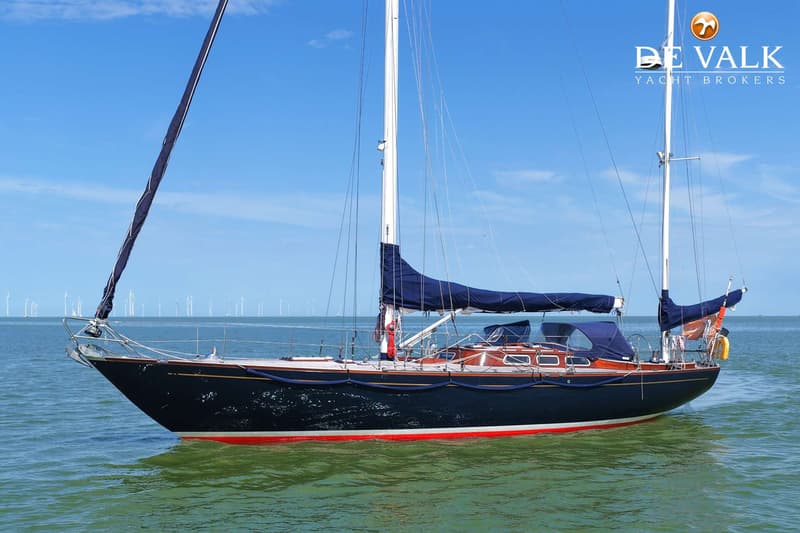 Een donkerblauwe zeilboot met rode en witte strepen op de romp vaart op groen water onder een blauwe lucht. — Huisman 45