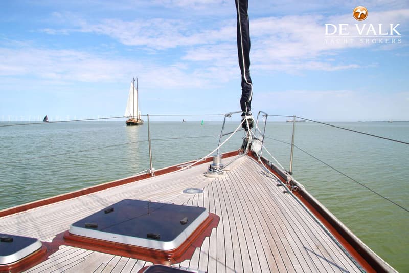 De foto toont het voordek van een zeilboot met een houten dek, uitkijkend over water met andere boten en windmolens aan de…