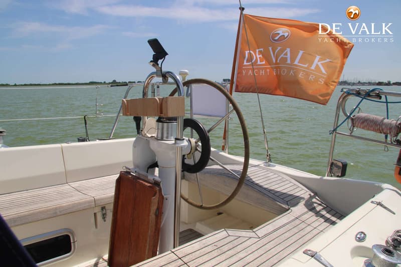 Een stuurstand met een houten stuurwiel en een oranje vlag met "DE VALK YACHT BROKERS" erop, op een boot met teakhouten dek.