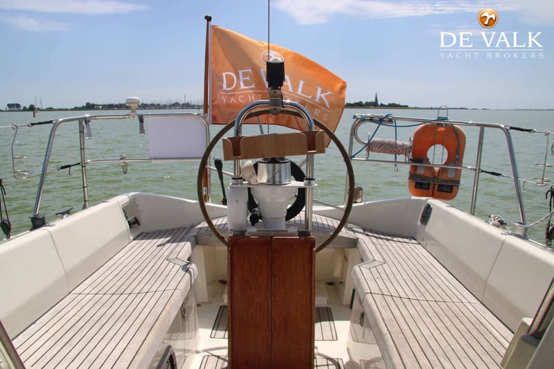Een achterzicht van een zeilboot met een stuurwiel, een oranje vlag en een reddingsboei, varend op water onder een blauwe…