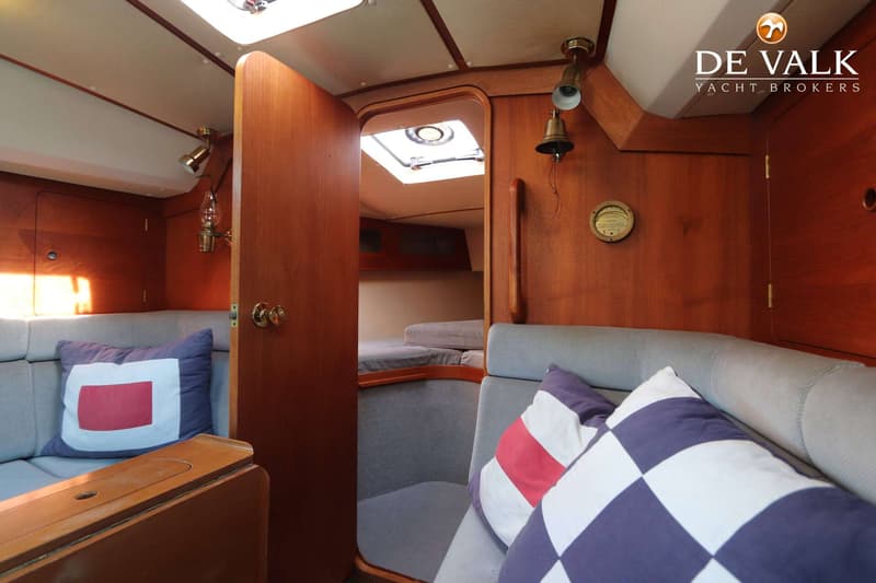 De foto toont een interieur van een boot met houten wanden, grijze banken met kussens, en een open deur naar een andere…