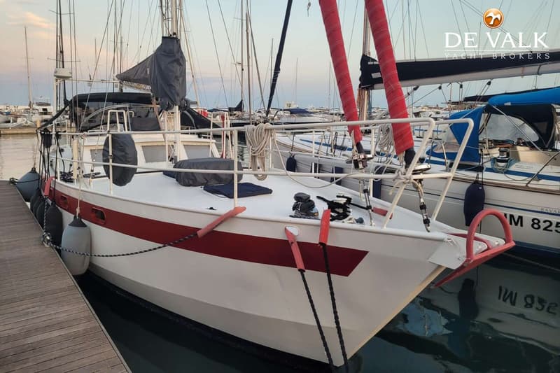 Een witte zeilboot met een rode streep op de romp ligt aangemeerd aan een houten steiger, met andere boten op de achtergrond.