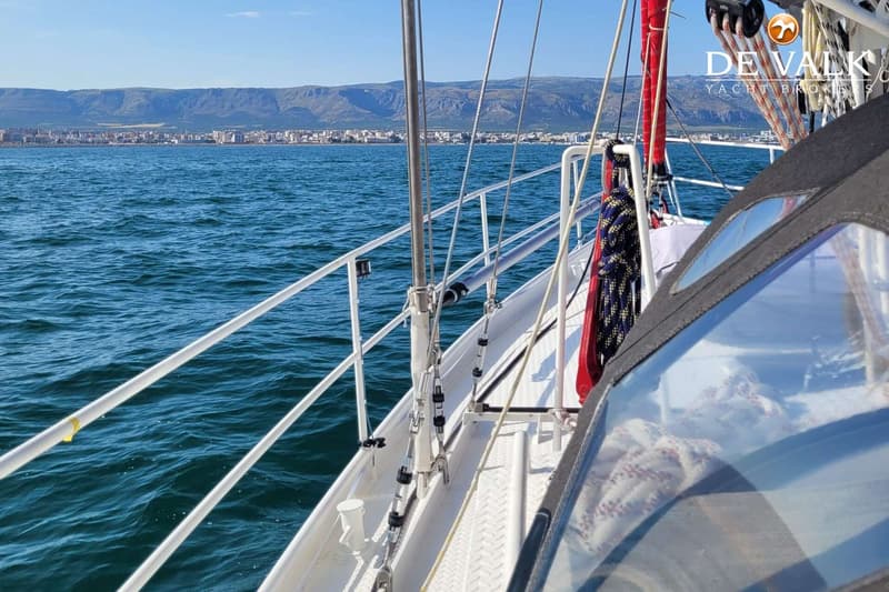 Een zeilboot vaart op blauw water met een stad en bergen op de achtergrond onder een heldere hemel. — Custom Built Sloop