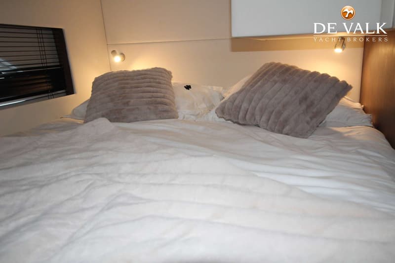 Een bed met wit beddengoed en twee grijze kussens, met een raam met jaloezieën en een lampje aan de muur.