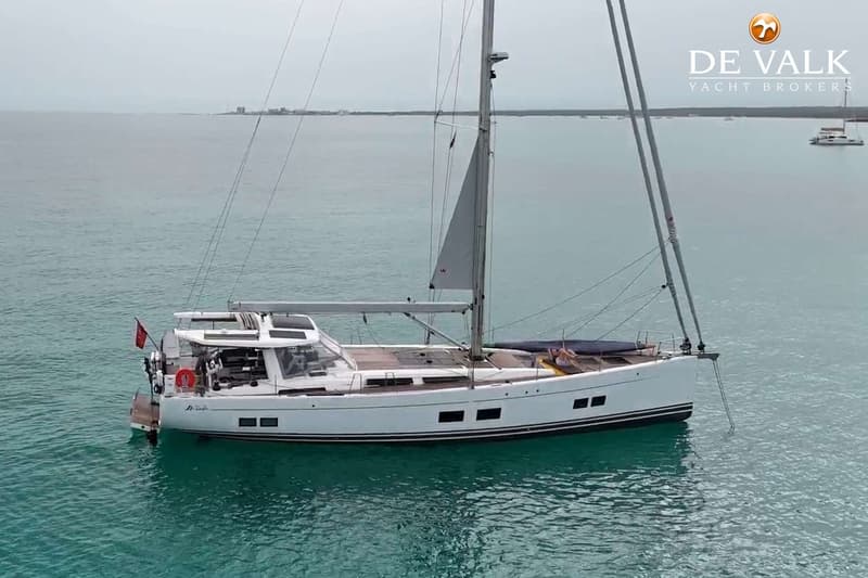 Een witte zeilboot met een opgerold zeil ligt voor anker in turquoise water, met een kustlijn op de achtergrond. — Hanse 588