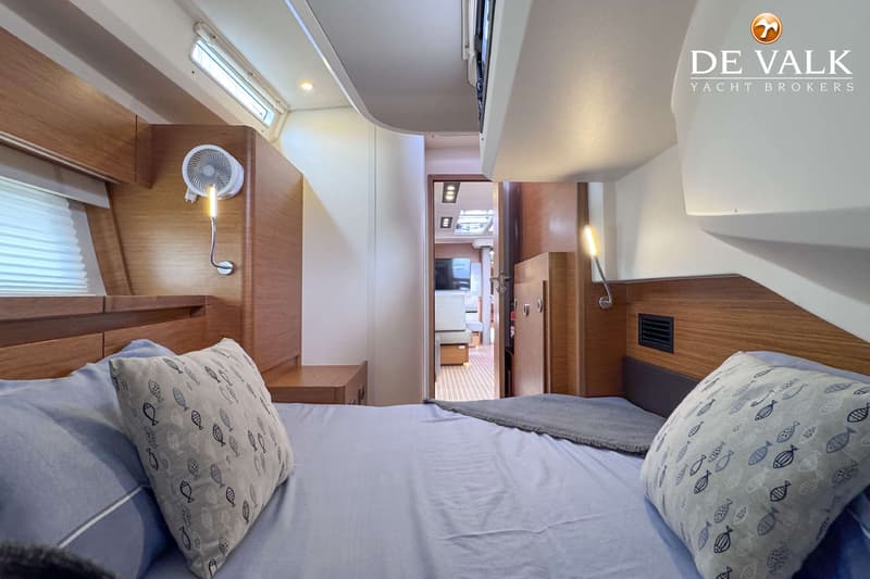 Een slaapkamer in een boot met een bed, kussens, een ventilator, lampen en een doorgang naar een andere ruimte. — Hanse 588