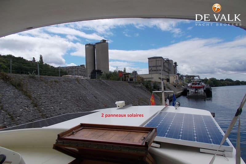 De foto toont het dek van een boot met twee zonnepanelen en een houten luik, met op de achtergrond een industrieel…