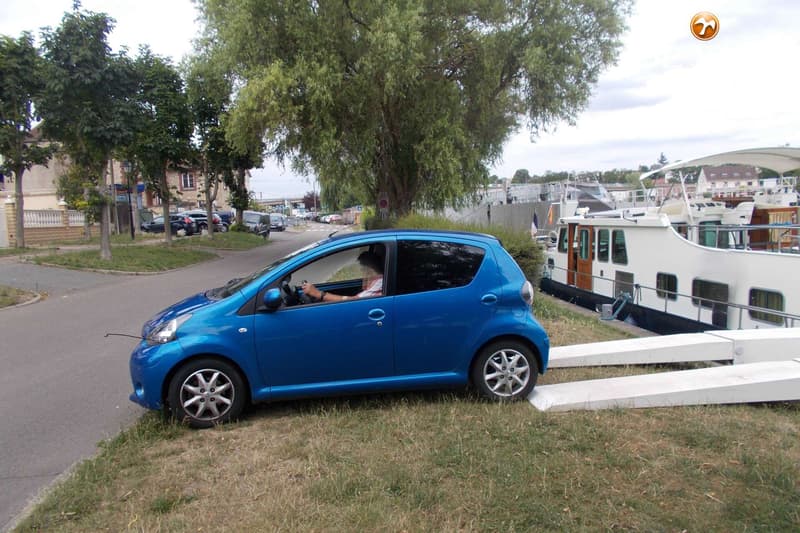 Een blauwe auto staat op een grasveld naast een kanaal met een witte boot en twee witte oprijplaten. — Tin Line 65