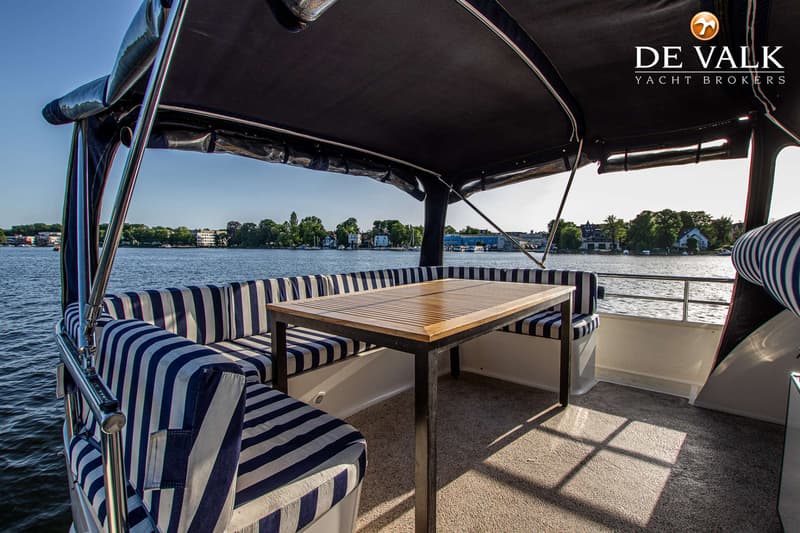 Een bootdek met een houten tafel, gestreepte kussens en een zwart zonnedak, met uitzicht op water en beboste oevers.